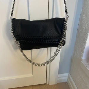 Svart axelväska med kedjedetaljer - Snygg svart axelväska i syntetmaterial med coola kedjedetaljer längs kanterna. Väskan har en stilren design och passar perfekt för en trendig look. Den har en lång kedjerem som gör den enkel att bära över axeln.