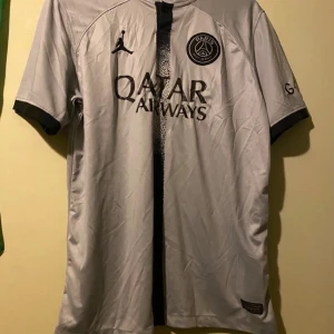 Grå fotbollströja från PSG - Snygg grå PSG-fotbollströja med korta ärmar och svart detalj i mitten. Tröjan har Jordan-logga och Qatar Airways-tryck på framsidan. Perfekt för fotbollsfans!