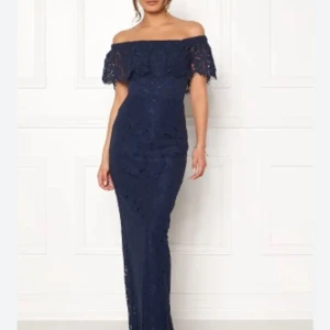 Marinblå spetsklänning - Elegant marinblå långklänning i spets med offshoulder-design. Klänningen har en vacker volangdetalj vid axlarna och en subtil slits baktill. Perfekt för festliga tillfällen.