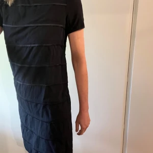 Svart Hugo Boss klänning - Snygg svart klänning från Hugo Boss. Korta ärmar,  horisontella detaljer som ger en stilren look samt guldfärgad dragkedja baktill.  Perfekt för en chic och minimalistisk stil.