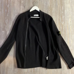 Svart ribbad jacka från Stone Island - Snygg svart ribbad jacka från Stone Island med dragkedja och långärmad design. Jackan har en broderad logotyp på ärmen som ger en stilren touch. Perfekt för en modern och avslappnad look.
