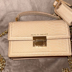 Beige handväska från Ideal of Sweden - Snygg beige handväska från Ideal of Sweden med krokodilpräglat mönster och gulddetaljer. Väskan har en praktisk kedjerem och ett elegant lås framtill.