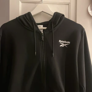 Svart hoodie från Reebok - Säljer en svart hoodie från Reebok med dragkedja och justerbar huva. Tröjan har vita logodetaljer på bröstet och längs ärmarna. Perfekt för en sportig look eller avslappnad stil.