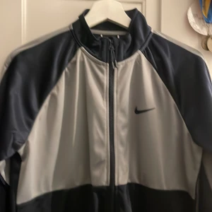 Svart och grå träningsjacka från Nike - Snygg träningsjacka från Nike i svart och grått med dragkedja framtill. Perfekt för sportiga aktiviteter eller som en stilren vardagsjacka. Den har långa ärmar och en klassisk passform. Byxorna finns också!