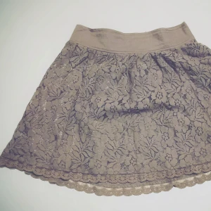 Beige spetskjol - Charmig beige kortkjol i spets med blommigt mönster. Kjolen har en elastisk midja för bekväm passform och en vacker volangkant som ger extra stil. Perfekt för en romantisk look.