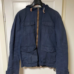 Mörkblå jacka från Selected - Säljer en mörkblå jacka från Selected med dragkedja och flera fickor framtill. Jackan har en huva och är långärmad, perfekt för kyligare dagar. Den är tillverkad i ett slitstarkt material och har en stilren design.