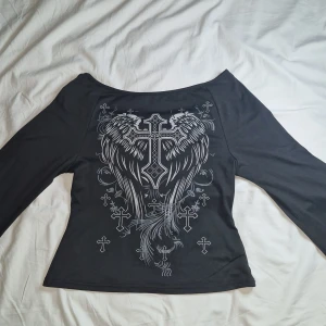 Svart långärmad topp med kors - Snygg svart långärmad topp med ett coolt tryck av kors och vingar på framsidan. Perfekt för en edgy look. Toppen har en normal passform och är tillverkad i ett mjukt material.