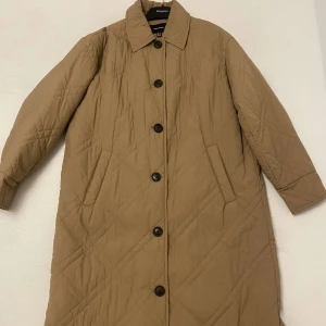 Beige quiltad kappa från Vero Moda - Säljer en stilren beige quiltad kappa från Vero Moda. Den har långa ärmar och knäpps med knappar framtill. Perfekt för kyligare dagar med sin långa och bekväma passform.