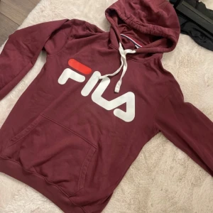 Vinröd hoodie från FILA - Säljer en snygg vinröd hoodie från FILA med stor logga på bröstet och mindre logga på ärmen. Tröjan har en praktisk magficka och justerbar huva med snören. Perfekt för en avslappnad stil. Alldrig  använd!