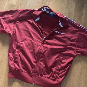 Röd kofta från Champion - Säljer en röd kofta från Champion med dragkedja och långa ärmar. Jackan har Champion-loggan på bröstet och längs kragen. Perfekt för en sportig look!