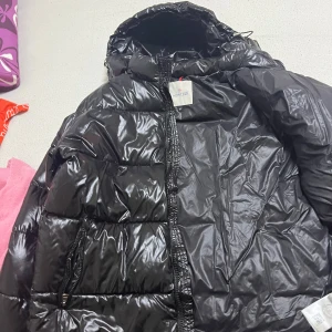 Svart Moncler puffer jacka  - Snygg svart pufferjacka från Moncler med glansig finish. Jackan har en dragkedja framtill och en praktisk huva. Perfekt för kyliga dagar med stil.