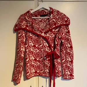 Röd och vit stickad kofta med knytband - Säljer en röd och vit stickad kofta med ett snyggt mönster. Koftan har knappar och knytbart band i midjan. 