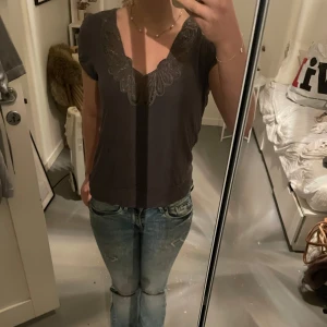 Grå topp från H&M med spetsdetaljer - Säljer en stilren grå topp från H&M med vackra spetsdetaljer vid halsringningen. Toppen har korta ärmar och en bekväm passform. Perfekt för en avslappnad men elegant look.