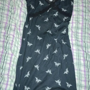 Svart klänning med fjärilsmönster - Snygg svart klänning med vita fjärilar och spetsdetaljer. Den har en v-ringad design med spetskant och ett fint knytband framtill. Perfekt för en unik och stilren look.