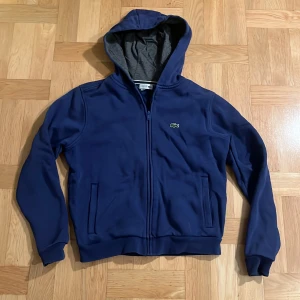 Blå hoodie från Lacoste - Säljer en snygg blå hoodie från Lacoste med dragkedja och huva. Den har långa ärmar och en liten krokodil-logga på bröstet. 