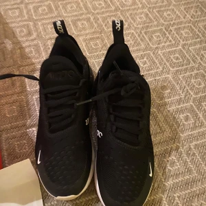 Svarta Nike Air Max 270 - Säljer ett par svarta Nike Air Max 270 med vit sula och synlig luftenhet i hälen. Skorna har snörning och Nike-logga i vitt. Perfekta för en sportig look.