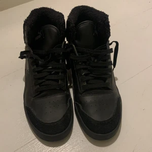 Svarta sneakers från Puma - Säljer ett par svarta sneakers från Puma med snörning och pälsfoder. Skorna har en hög design och är perfekta för kyligare väder.