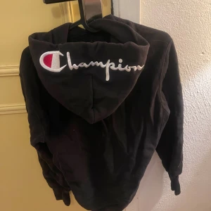 Svart hoodie från Champion - Säljer en svart hoodie från Champion med stor logga på ryggen. Perfekt för en avslappnad stil. Den har en klassisk huva och långa ärmar.