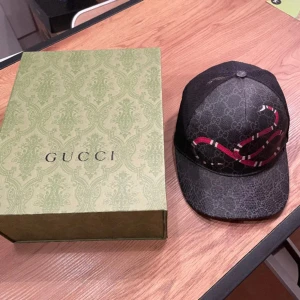Svart keps med ormmönster från Gucci - Snygg svart keps från Gucci med ett rött ormmönster. Kepsen har ett klassiskt Gucci-mönster i bakgrunden och justerbar passform. Perfekt för att ge din outfit en lyxig touch. Boxen finns inte längre av pågrund av en flytt annars e den i underbar skick❤️ Stl M men passar dom andra stl oxå såklart