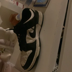 Svarta och vita Nike sneakers - Snygga svarta och vita Nike sneakers med klassisk design. Skorna har snörning och en bekväm passform, perfekt för vardagsbruk. Den ikoniska Nike-loggan syns tydligt på sidan.