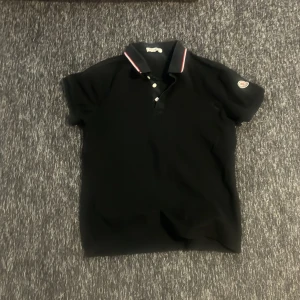 Svart kortärmad pikétröja från Moncler - Snygg svart pikétröja från Moncler med korta ärmar och klassisk krage. Tröjan har knappar framtill och en diskret logga på ärmen. Perfekt för en stilren look.