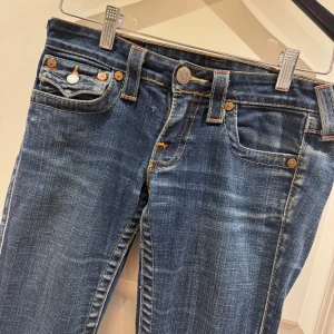 Blå jeans från True Religion - Snygga blå jeans från True Religion med klassisk femficksdesign och kontrastsömmar. De har en bootcut-stil och är dekorerade med märkets ikoniska broderade fickor bak. Perfekta för en avslappnad look. Låg midja. Midjemåttet är ca 40 och innerbenslängd 80. De har lite slitningar längst ner 