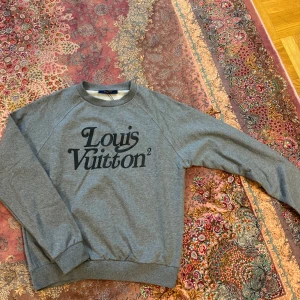 Grå sweatshirt från Louis Vuitton - Snygg grå sweatshirt från Louis Vuitton med svart logotyptryck på både fram- och baksidan. Tröjan har långa ärmar och rund halsringning. Perfekt för en stilren och avslappnad look.