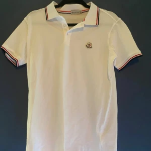 pikétröja från Moncler - Snygg vit pikétröja från Moncler med korta ärmar och klassisk krage. Tröjan har röd och blå randdetalj på krage och ärmslut samt Moncler-logga på bröstet. Perfekt för en stilren look.