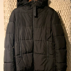 Svart pufferjacka från Bershka - Snygg svart pufferjacka från Bershka med dragkedja och huva. Perfekt för kyliga dagar med sin quiltade design och praktiska fickor. Jackan har långa ärmar och en stilren look.