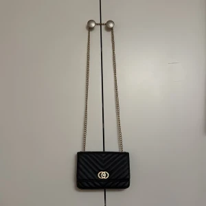 Svart väska  - Snygg svart axelväska i quiltat mönster med elegant guldkedja och dekorativt spänne framtill. Perfekt för att ge en lyxig touch till din outfit.