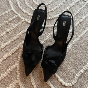 Svarta klackskor med blomdetalj från Zara - Eleganta svarta klackskor från Zara med spetsig tå och en vacker blomdetalj på ovansidan. Skorna har en smal rem runt hälen och en stilren klack som ger en sofistikerad look.