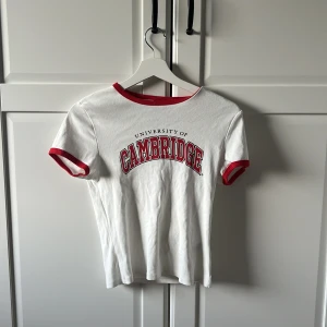 Vit t-shirt från H&M Divided med Cambridge-tryck - Snygg vit t-shirt från H&M Divided med rött Cambridge-tryck. Den har röda kanter vid hals och ärmar. Passar jättebra med en kjol eller ett par snygga jeans. Perfekt för våren och sommaren!