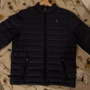 Ralph Lauren jacka - Snygg blå pufferjacka från Ralph Lauren med röd insida och dragkedja. Jackan är i nytt skick och oanvänd, köptes från Kidsbrandstore för 2499 kr, jackan har en skön och bekväm insida.  Perfekt för kyliga dagar, tveka inte för att diskutera om ett bra pris. Den är stilren och passar för vintern 