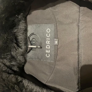 Svart dunjacka från Cedrico - Snygg svart dunjacka från Cedrico med pälskantad huva och dragkedja framtill. Jackan har långa ärmar och praktiska fickor med dragkedjor. Perfekt för kalla dagar. Köpt från Johnells, har en liten defekt. 