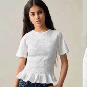 Vit blus med volangdetalj - Säljer en söt vit blus med smockdetaljer och volang vid nederkanten. Blusen har korta ärmar och en rund halsringning. Perfekt för en stilren look. Nästan som ny, använda 5/6 gånger 