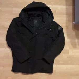 Vinterjacka - Säljer en svart dunjacka från Canada Goose med huva och broderad logga på ärmen. Jackan har en stilren design med knappar och dragkedja framtill. Perfekt för kalla dagar.