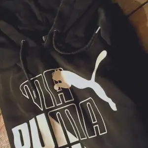 Säljer en snygg svart hoodie från Puma. Den har en stor vit logga på framsidan och justerbara snören i huvan. Perfekt för en avslappnad stil eller träning. Passar bra till både jeans och träningsbyxor!