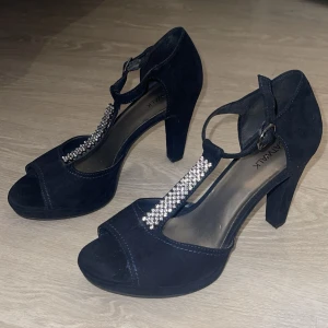 Svarta klackskor med strassdetaljer - Säljer ett par eleganta svarta klackskor från Catwalk. De är i mocka med en öppen tå och har en snygg rem med strassdetaljer som ger en glittrig touch. Perfekta för festliga tillfällen! Skorna har en bekväm klack och justerbar rem runt ankeln.