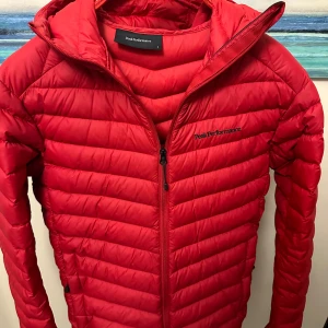 Röd dunjacka från Peak Performance - Säljer en snygg och varm röd dunjacka från Peak Performance. Jackan har en dragkedja framtill och är perfekt för kyliga dagar. Den är långärmad och har en stilren design med en huva för extra skydd mot väder och vind.