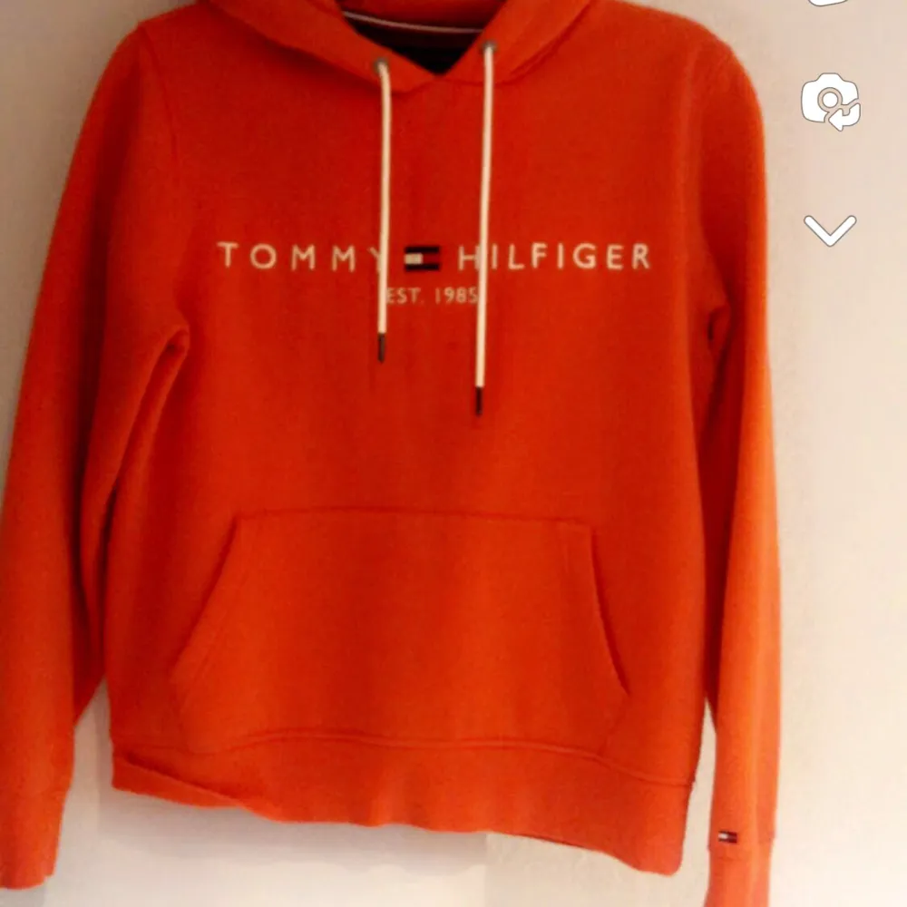 Säljer en snygg orange hoodie från Tommy Hilfiger med deras klassiska logga på bröstet. Den har en känguruficka och vita snören i huvan. Perfekt för en casual look eller när du vill ha något bekvämt men stiligt. Passar både till vardags och chillkvällar.. Neuletakit & Villapaidat.