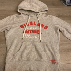 Grå hoodie från H&M - Säljer en grå hoodie från H&M:s L.O.G.G.-kollektion. Den har en stor ficka fram och texten 'Highland Cinemas Matinee Classic' i rött och vitt på bröstet. Perfekt för en avslappnad stil eller mysiga dagar hemma. 🩶