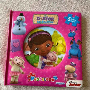 Doktor McStuffins Pusselbok - En färgglad och rolig pusselbok med Doktor McStuffins från Disney Junior. Boken innehåller 5 olika pussel som barn kan lägga medan de följer med på äventyr med Doktorn och hennes vänner. Perfekt för små barn som älskar att läsa och pussla samtidigt. Boken har en härlig rosa färg med bilder av karaktärerna på framsidan.