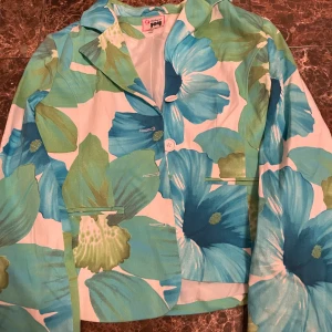 Blommig kavaj från Joy - Snygg kavaj med stort blommönster i blått, grönt och vitt. Perfekt för att liva upp garderoben med en färgglad touch. Kavajen har långa ärmar och knappar framtill. Köpt på flash aldrig använt tidigare 