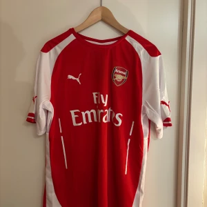 Röd Arsenal fotbollströja från Puma - Säljer en röd Arsenal fotbollströja från Puma med vita detaljer och korta ärmar. Tröjan har Arsenal-loggan och Fly Emirates-tryck på framsidan. Perfekt för alla Arsenal-fans! 🏆