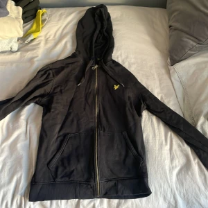Svart huvtröja från Lyle & Scott - Säljer en svart huvtröja från Lyle & Scott med dragkedja och fickor framtill. Tröjan har en gul broderad logga på bröstet och ribbade muddar vid ärmslut och nederkant. Perfekt för en avslappnad stil.