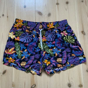 Färgglada blommiga shorts från Vans - Säljer ett par färgglada shorts från Vans med ett tropiskt blommönster i blått, rosa, grönt och gult. Shortsen har en bekväm passform med snörning i midjan. Står att det är storlek M men skulle säga att den passar mer som S. 