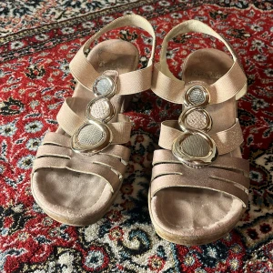 Beige sandaler med klack från Rieker - Snygga beige sandaler från Rieker med klack och dekorativa metallringar på ovansidan. Perfekta för sommarens alla tillfällen. Sandalerna har en bekväm passform och elastiska remmar för enkel på- och avtagning.