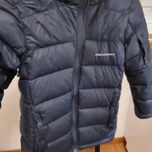 Mörkblå dunjacka från Peak Performance - Säljer en mörkblå dunjacka från Peak Performance. Jackan har en dragkedja framtill och en praktisk huva. Perfekt för kyliga dagar med sin vadderade design. Dragkedja i en fickan fram funkar inte men annars inga fel. Behöver ev en tvätt 