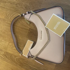 Rosa axelväska från Michael Kors - Snygg rosa axelväska från Michael Kors i skinn med guldfärgad kedjedetalj och dragkedja. Perfekt för att ge en touch av lyx till din outfit. Väskan har en stilren design och är rymlig nog för dina nödvändigheter. Köpt men aldrig använd, nypris: 538$ motsvarande ca 6000kr (den ser beige ut på bild men är rosa) pris kan diskuteras vid snabb affär 