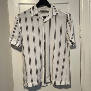 Vit och svart randig skjorta från MNG - Säljer en stilren vit skjorta med svarta ränder från MNG. Skjortan har korta ärmar och knappar framtill. Knappt använd, mycket bra skick. Perfekt till sommaren!